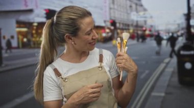 Makyaj fırçalarını elinde tutarken gülümseyen kadın, şehrin işlek caddelerinin köşesinde yaratıcı neşe.