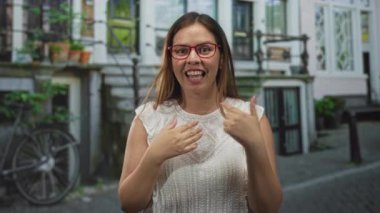 Kadın, bisiklet ve inşaat cephesi ile kaldırım taşı caddesinde göğsüne dokunurken ve omuzlarını silkerken kamerayı işaret ediyor..