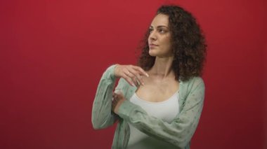 Kadın, yeşil tişört ve beyaz tankla kırmızı stüdyoda omuz ve el göstererek parmağını yukarı kaldırıyor; özgüven tutkusu.