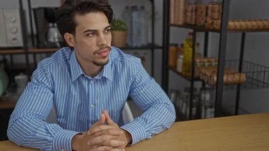 Ofiste oturan genç adam, çizgili gömlek giymiş, elleri masaya yapıştırılmış. Mutfak aletleri rafları ve kavanozlarla çevrili düşünceli görünüyor..