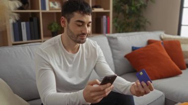 Oturma odasındaki kanepede otururken elinde akıllı telefon ve kredi kartı tutan genç adam online alışveriş öneriyor.