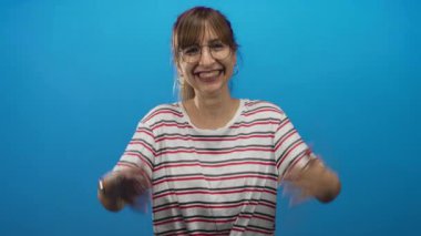 Mavi stüdyoda geniş ve açık avuç içlerine gülümseyen çizgili gömlek giymiş, kollarını uzatmış ve görünür kollarıyla öne doğru uzanan bir kadın..