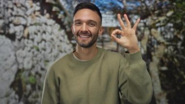 Parlak yeşil bir parkta iki eliyle el kol hareketi yapan genç adam, açık havada mutlu bir şekilde gülümsüyor sakin bir ortamda bereketli doğayla çevrili..