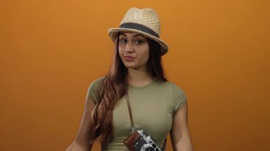 Şapkalı kadın, canlı turuncu arkaplana karşı kamerayla güven içinde poz veriyor. Huzur ve macera ruhunu ifade ediyor. Genç gezginlerin özünü somutlaştırıyor..