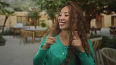 Kadın açık hava restoranında terası işaret ediyor, kırmızı tırnakları ve dalgalı kıvırcık saçlarıyla gülümsüyor; kaygısız neşe.