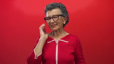 Gri saçlı, kırmızı elbiseli, akıllı telefonlu, kırmızı arka planda konuşan ve gülümseyen bir kadın..