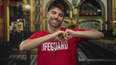 Kırmızı cankurtaran gömleği giyen adam elleriyle kalp şekli oluşturuyor ve kilisenin içinde geniş bir gülümseme sergiliyor; şefkat..