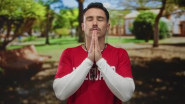 Kırmızı gömlekli genç İspanyol cankurtaran yeşil ağaçlar ve parlak güneşli bir çevre ile çevrili, huzurlu bir açık hava parkı ortamında gözleri kapalı meditasyon yapıyor..