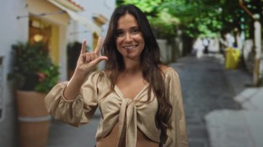 Seni seviyorum derken gülümseyen bir kadın güneş ışığıyla aydınlanmış bir caddede çiçek saksısı ve kaldırımlı bir kaldırım taşı ile el işareti yapıyor..