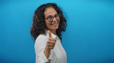 Kıvırcık saçlı, gülümseyen ve mavi arka planda baş parmak kaldıran neşeli ve izole bir ortamda mutluluk ve kendine güven saçan bir kadın..