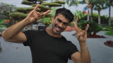 Kaya boynuzları yapan genç İspanyol adam, yeşil ağaçların ve fıskiyelerin yanındaki sokakta elleri ve gülümseyerek hareket ediyor. Rock metal eğlencesi..