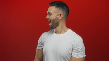 Young hispanic man in white shirt excitedly reacts against vibrant red background suggesting surprise and joy