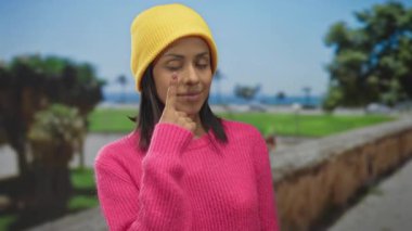 Pembe kazaklı ve sarı şapkalı genç bir kadın güneşli bir parkta gözlerini işaret ediyor. Ağaçlar gür ve sakin bir geçmişi var. Huzur ve tefekküre önem veriyor..