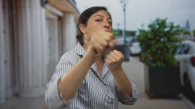 Latin kökenli orta yaşlı bir kadın, şehir sokağında gündüz vakti yumruklarını kaldırarak kameraya yumruk atıyor..