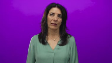 Stüdyoda yeşil bluz ve inci kolye giyen, mor arka planda parmak ucunda silah tutan İspanyol bir kadın hayal kırıklığı..