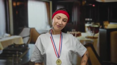 Madalyon takan bir kadın restoranda kendine güvenerek ayakta duruyor. Kırmızı bir saç bandıyla şık bir yemek geçmişine karşı günlük giysiler içinde atletik başarılar sergiliyor..