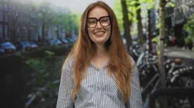 Kırmızı saçlı ve gözlüklü gülümseyen kadın, güneş ışığıyla aydınlanan caddede kamerayı işaret ediyor..