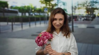 Şehir caddesinde çiçeklerle gülümseyen genç bir kadın, ağaçlarla ve bulanık binalarla huzurlu bir şehir ortamını öneriyor, mutluluk ve sükuneti somutlaştırıyor..