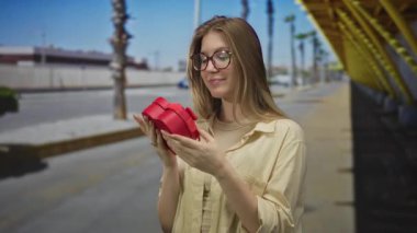 Kadın, sarı iskele ve palmiye ağaçlarının yanında güneşli şehir sokağında iki eliyle parlak kırmızı bir hediye kutusu tutuyor..