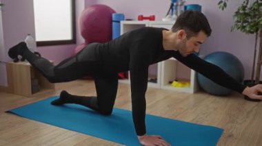 Spor salonundaki minderde yoga yapan, arka planda spor aletleri olan bir adam, kapalı alanda egzersiz yaparken denge ve kuvvete odaklanıyor..