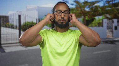 Evin girişinde parmaklarıyla kulaklarını tıkayan adam, parlak yeşil gömlek ve gözlük takıyor, yüksek sese tepki olarak gözler kapalı; gürültü rahatsız edici.