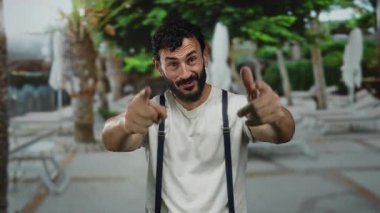 Sakallı İspanyol bir adam güneşli bir sokakta arka planda palmiye ağaçları olan beyaz bir gömlek ve pantolon askısı takarak coşkuyla el kol hareketi yapıyor..