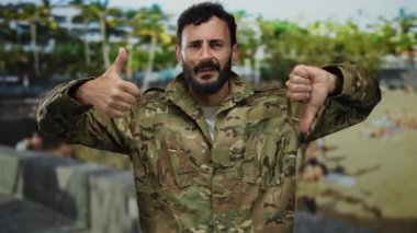 Sakallı, kamuflaj üniformalı bir asker yeşil bir parkta arka planda ağaçlarla dikilirken ve dışarıda karışık duygular gösterirken baş parmağıyla işaret veriyor..