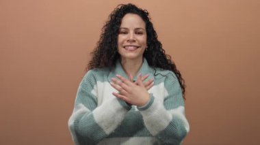 Çizgili süveter giyen bir kadın izole edilmiş kahverengi bir duvar arkasına sıcak bir şekilde gülümsüyor, açık jestler, kıvırcık saçlar ve gündelik tarzda dostça ve davetkar davranışlar sergiliyor..