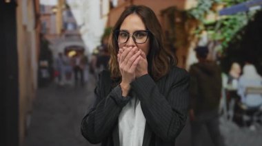 Kalabalık bir şehir caddesinde gözlüklü İspanyol kadın, bulanık insanlar ve kentsel çevre ile çevrili ağzı şaşkınlıkla kaplayarak, canlı bir şehir manzarası yaratıyor..