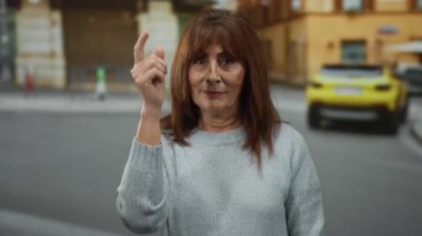 Üst düzey İspanyol kadın süveter giyerek şehir caddesinde dikiliyor şehir arka planında bulanık bir jest yapıyor ve arka planda sarı bir araba gün ışığının altında park halinde bekliyor.