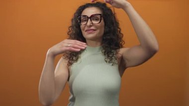 Kıvırcık saçlı, gözlüklü, yüzü turuncu stüdyo duvarına karşı elleri olan bir kadın..