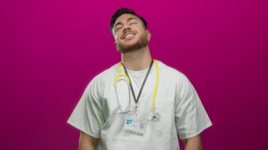 Steteskop ve sakallı, gülümseyen genç erkek doktor canlı pembe bir arka plana karşı duruyor, tıbbi bir ortamda profesyonel bakım ve neşeli bir tavır yakalıyor..