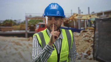 Mavi kasketli ve çelik yelekli bir adam, çizgili gömlek, parmak dudağa sessizlik için inşaat alanında dikkat..