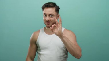 Beyaz askılı sakallı genç adam yeşil bir arka plana gülümseyen, dışa dönük, dışa dönük yüz ifadelerini sergileyen iyi bir işaret veriyor..