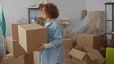 Parlak bir oturma odasında kutuları açan kadın, yeni dairesinde sayısız karton kutu ile çevrili, ev içine taşınırken neşe ve başarı ifade ediyor.