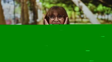 İspanyol kıdemli bir kadın, doğa ile çevrili bir parkta neşeyle gülümsüyor, gözlük takıyor ve kareli bir gömlek giyiyor, yeşil yapraklı ağaçların ve güneş ışığının arasında mutluluk ve sıcaklığı ifade ediyor..