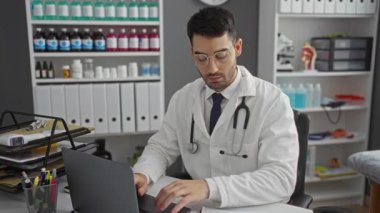 Beyaz önlüklü genç erkek doktor, hastane odasındaki bir masada çalışıyor. Raflarda tıbbi malzemeler var. Evrak işlerine odaklanmış ve dizüstü bilgisayar kullanıyor..