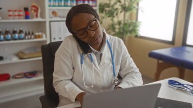 Doktor, hastane ofisinde steteskopu görünür olan telefon ve dizüstü bilgisayarla notlar yazıyor. Parlak bir klinik ortamında tıbbi malzemelerle çevrili..