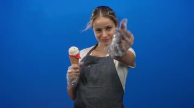 Gülümseyen ve eldivenli el ve kot önlüklü bir dondurma külahı sunan bir kadın stüdyoda mavi arka plana karşı, neşeli bir karşılama..