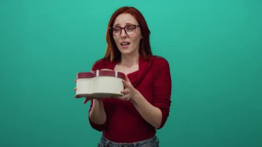Kızıl saçlı kadın yeşil arka planda bir hediye kutusu tutuyor, neşe ve heyecanı ifade ediyor, günlük kırmızı süveter ve neşeli bir tavırla gözlük giyiyor..