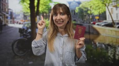 Kadın, güneşli bir şehir sokağında park halindeki bisikletli bir kanalda pasaportu tutarken yukarı doğru işaret ediyor; seyahat heyecanı.