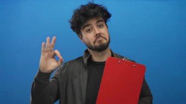 Sakallı genç adam iyi bir jest yapıyor ve kırmızı panoyu mavi arka planda tutuyor. Günlük kıyafetlerle güven ve onayı ifade ediyor..