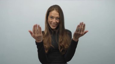 Avuç içi açık gülümseyen bir kadın soluk gri arka planı ve uzun kahverengi saçları ile stüdyoda jest yapmaya davet ediyor; sıcak davetli sevinç.