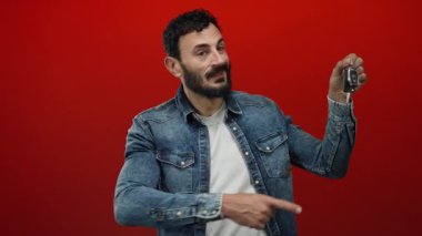 Kot ceketli İspanyol adam heyecan verici bir şekilde elinde kırmızı arka planlı araba anahtarı tutuyor. Promosyon amaçlı dinamik ve canlı bir atmosfer yaratıyor..