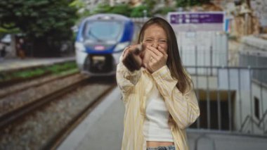 Tren istasyonundaki tren istasyonunda eli ağzı kapatırken genç İspanyol kadının parmağı kamerayı işaret ediyor; neşeli kahkahalar.
