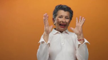 Beyaz tişörtlü, gri saçlı yaşlı bir kadın turuncu arka planda şaşkınlık yaratıyor. Duygularını, yaşını ve ifadesini canlı ve çekici bir şekilde gösteriyor..