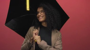 Şemsiye altında kırmızı arka planda gülümseyen, neşeli ifadeler ve şık kıyafetler sergileyen bir kadın..