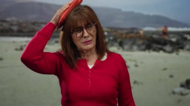 Kırmızı tişörtlü İspanyol son sınıf öğrencisi, puslu bir günde arka planda bulanık okyanus ve kayalarla deniz kenarındaki plajda dikilirken buz torbasını uyguluyor..