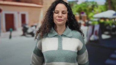 Genç İspanyol kadın güneşli bir caddede iki eliyle kaya jesti yaparken gülümsüyor, gözlük takıyor ve çizgili bir kazak giyiyor..