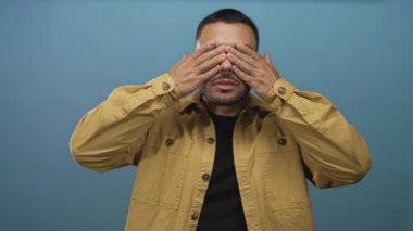 Hardal ceketli genç İspanyol adam mavi stüdyo duvarına karşı elleri ile gözlerini kapar hüzünlü poz; neşe dolu kaprisli kaprisli neşe.